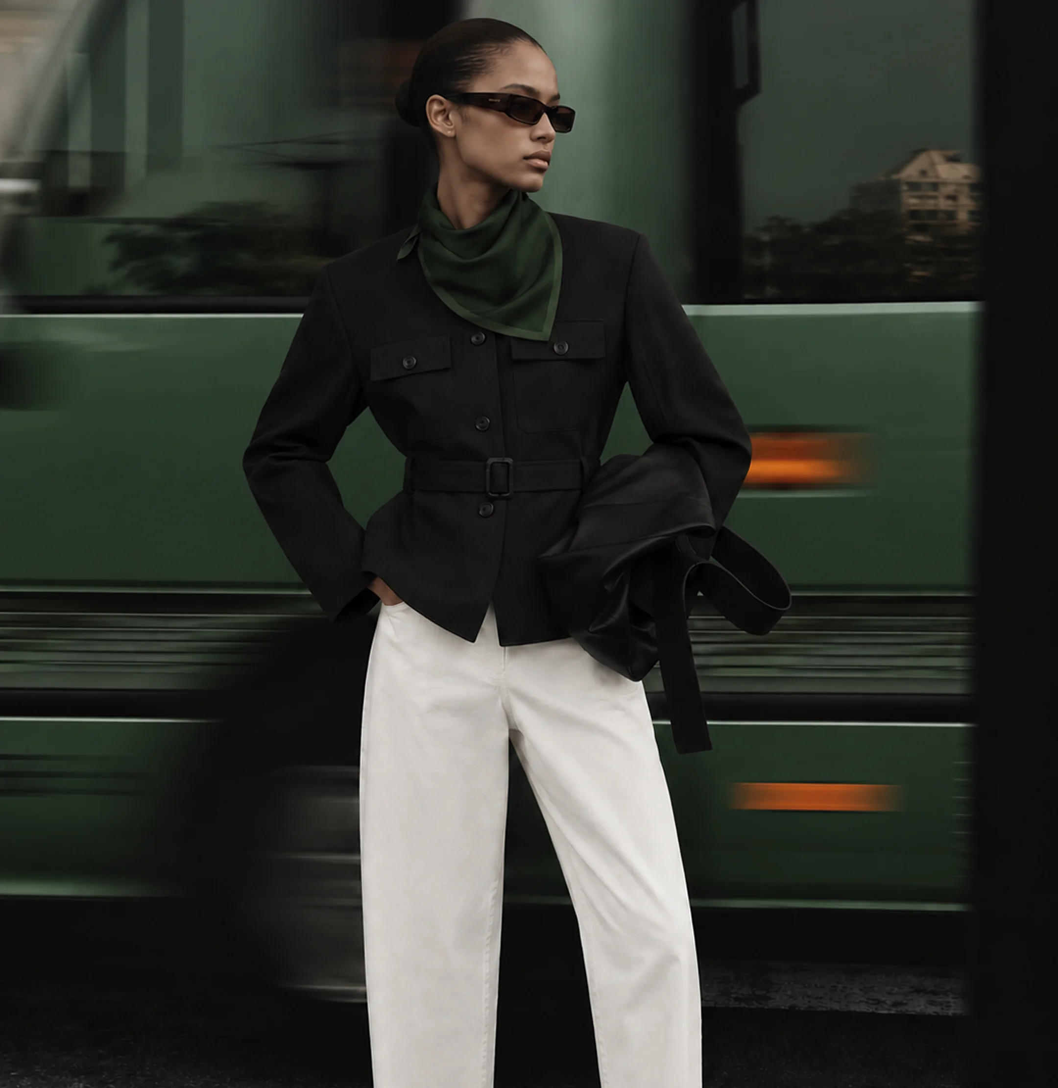 Woman in a black belted jacket standing in front of a moving bus
