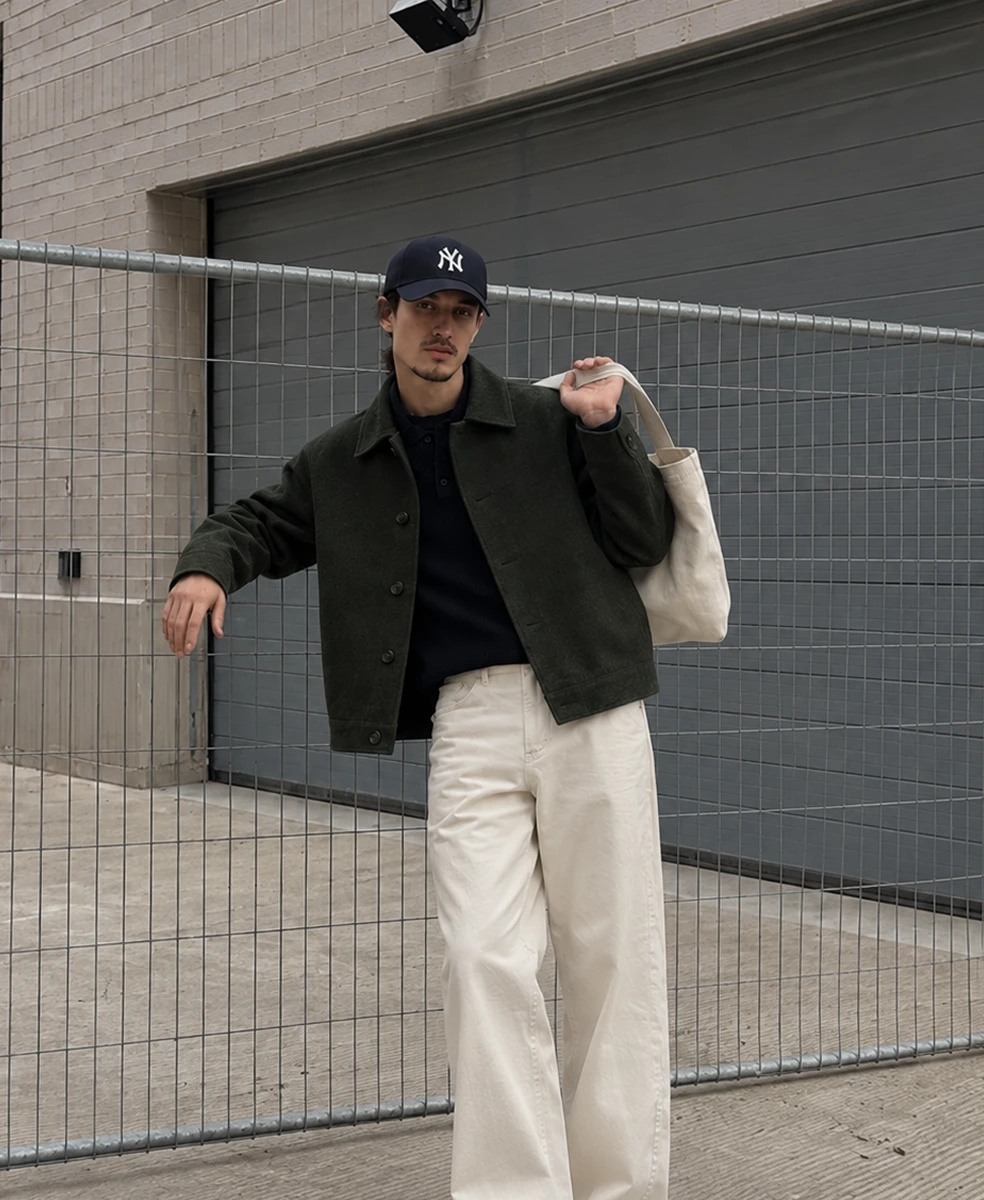 Man in a navy cap and dark green jacket with cream trousers carrying a tote bag by a metal fence