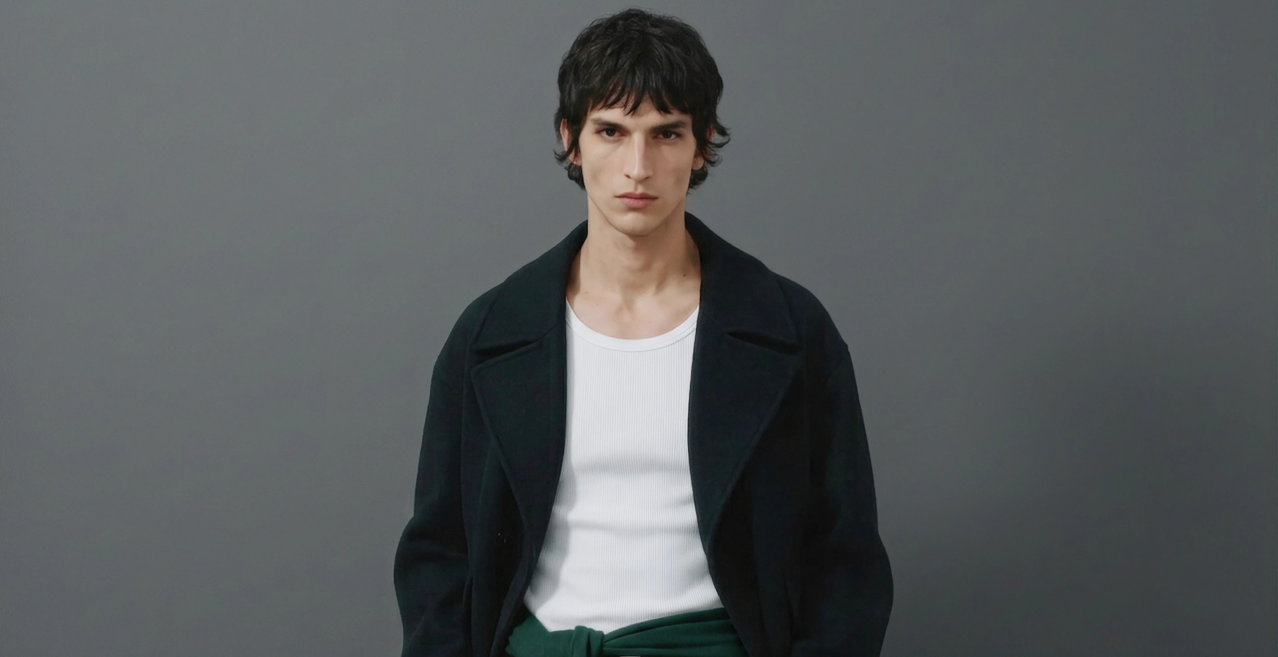Man in a dark navy coat over a white ribbed tank top with a green sweater tied at the waist